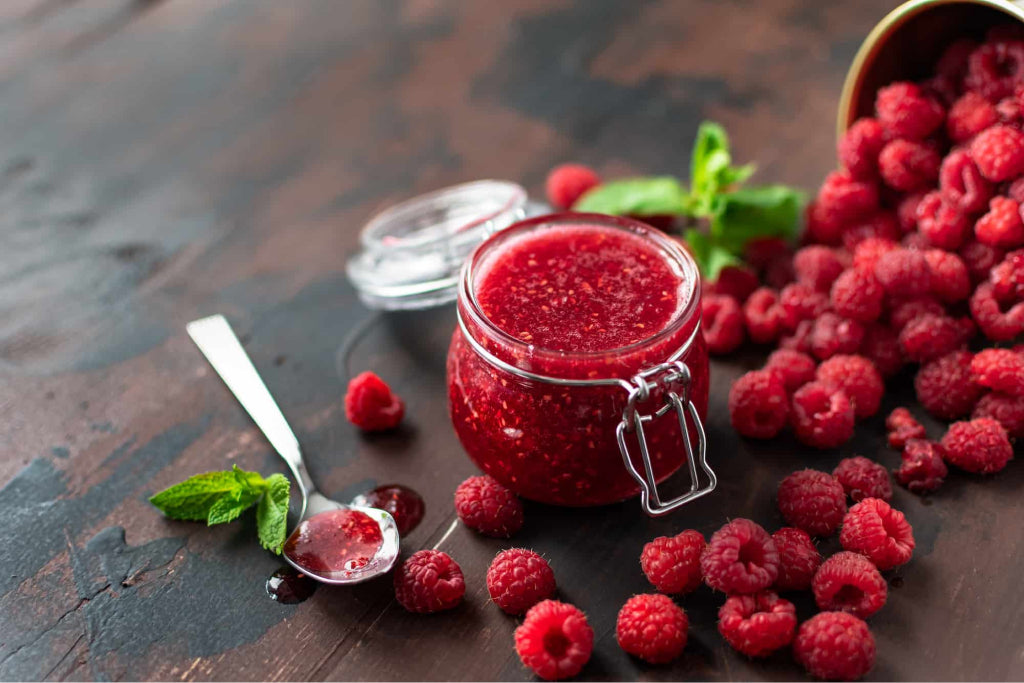 a jar of low carb raspberry jam on a kitchen counter with wild raspberries spread around. 