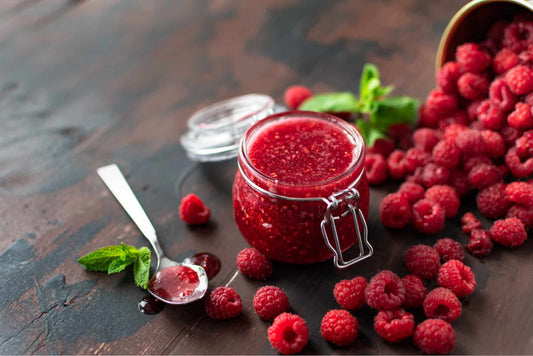 a jar of low carb raspberry jam on a kitchen counter with wild raspberries spread around. 