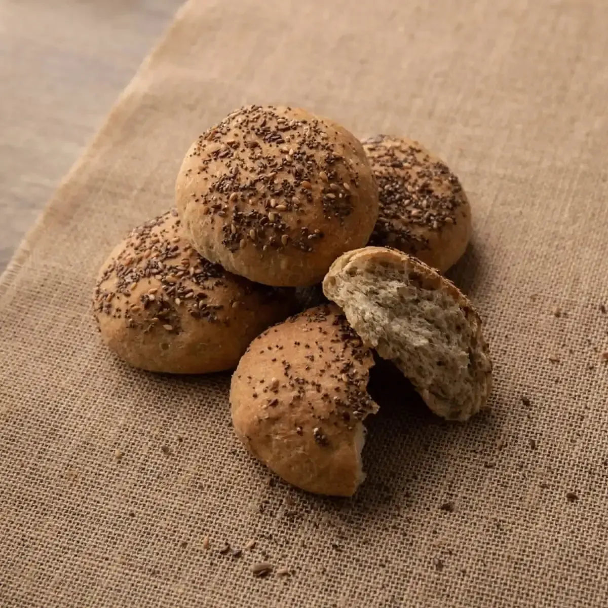 Rustic bread rolls with flax seeds on burlap, one broken open to show soft interior.