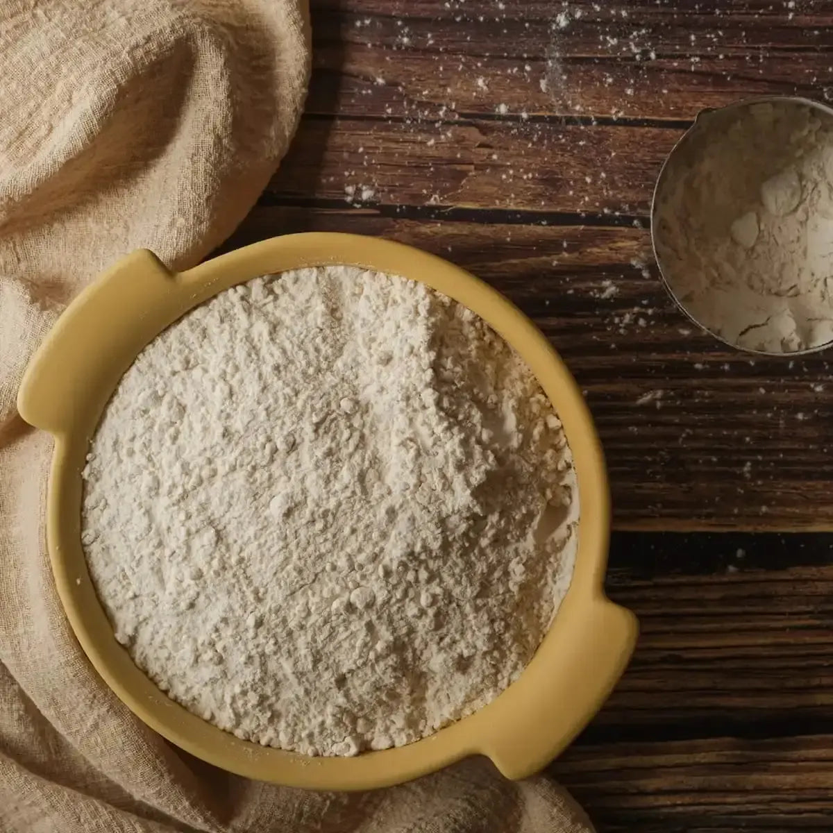 A yellow bowl of white flour on a rustic wooden table with a beige cloth.