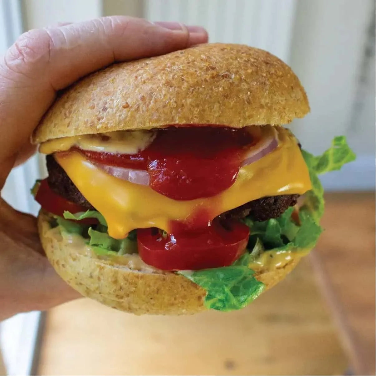Hand holding a keto-friendly burger with a brown bun, melted cheese, lettuce, tomato, onion, and ketchup.
