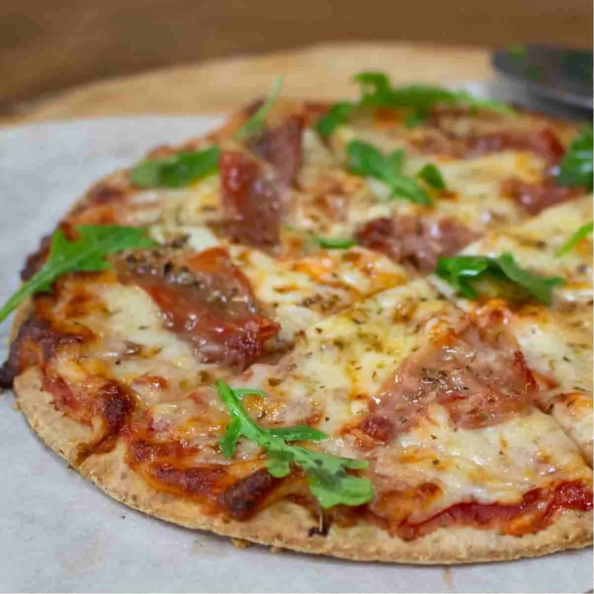 Golden-crusted keto pizza with melted cheese, red sauce, pepperoni, and fresh arugula on parchment paper.