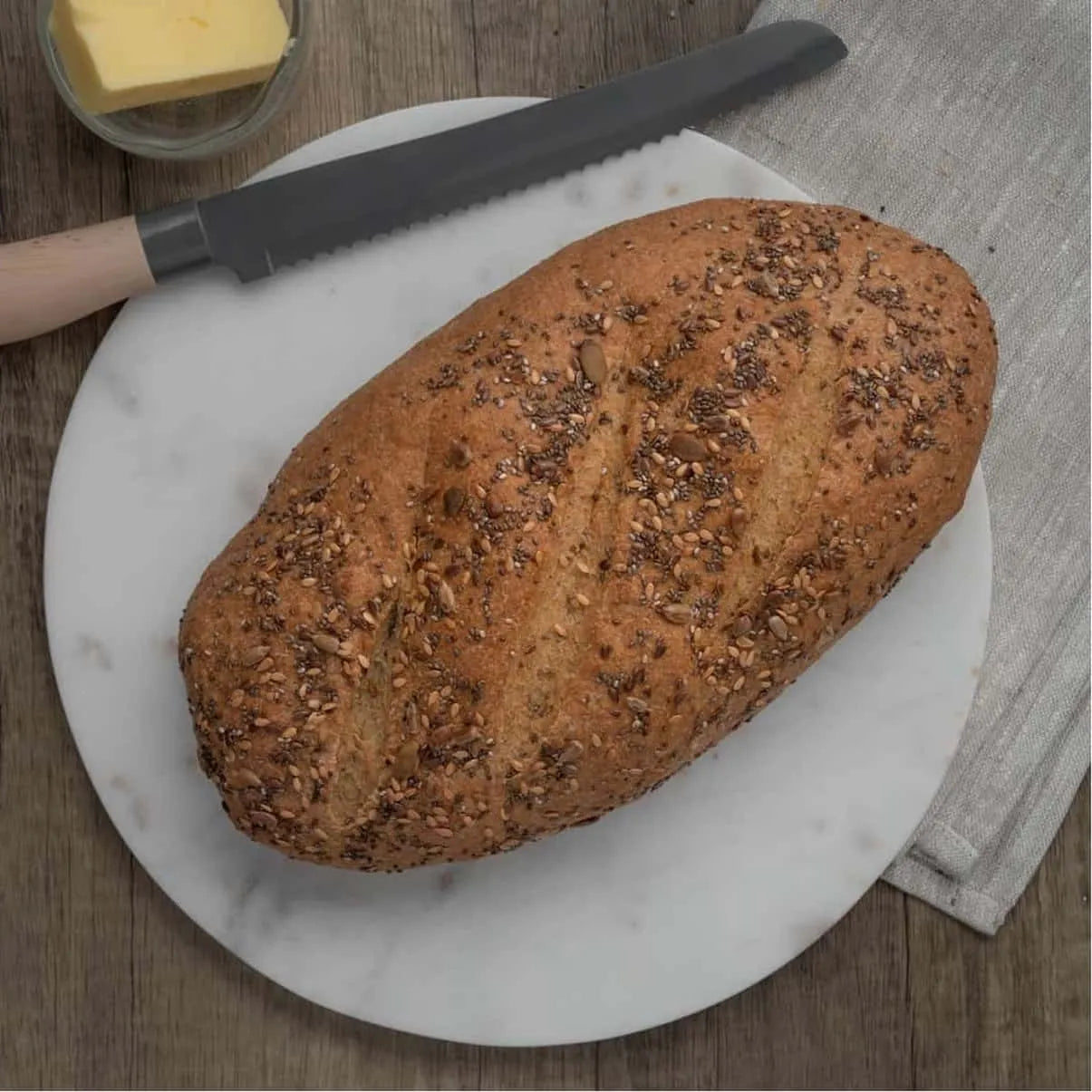 Golden brown seeded bloomer bread on a white marble board with a bread knife and butter.