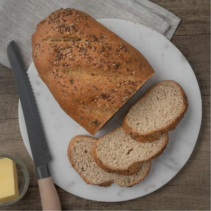 Seeded low-carb bloomer bread, partially sliced, on a white marble board with a bread knife and butter.