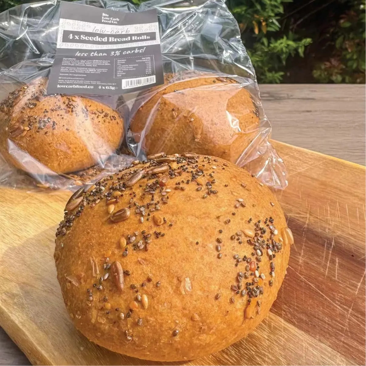 Golden brown keto seeded bread rolls, one unpackaged on a wooden board, two in clear packaging.