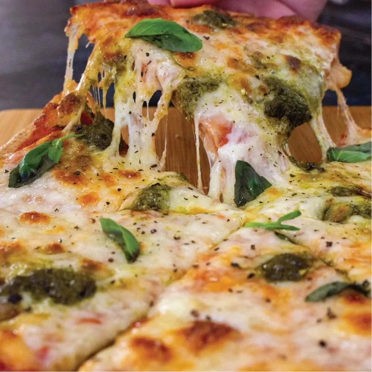 A hand lifts a slice of hot, cheesy keto pizza with pesto and fresh basil from a wooden board.