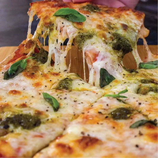 A hand lifts a slice of hot, cheesy keto pizza with pesto and fresh basil from a wooden board.