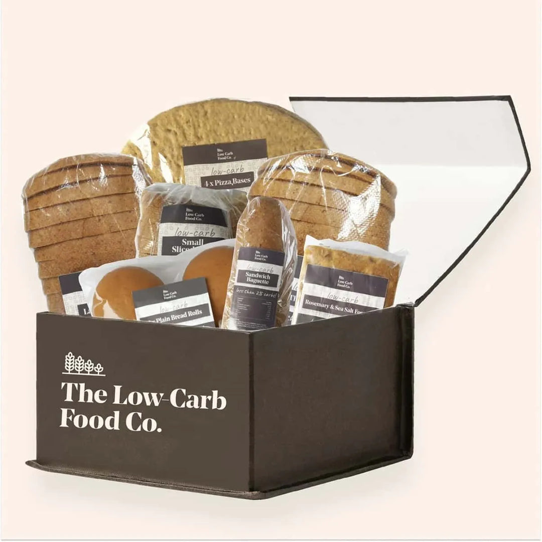 Open brown box filled with various packaged low-carb breads: sliced loaves, rolls, baguette, focaccia, and pizza bases.