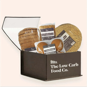 Open black box filled with various packaged low-carb breads: sliced loaf, focaccia, rolls, and pizza bases.